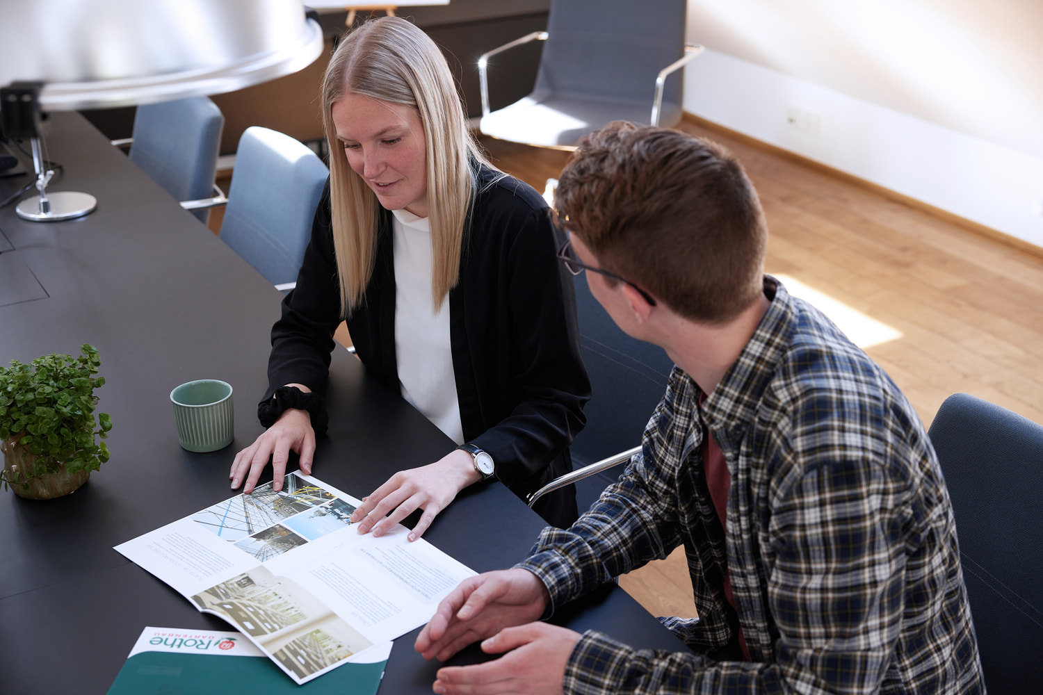 Two colleagues are sitting at a table and looking at brochures while pointing and talking.