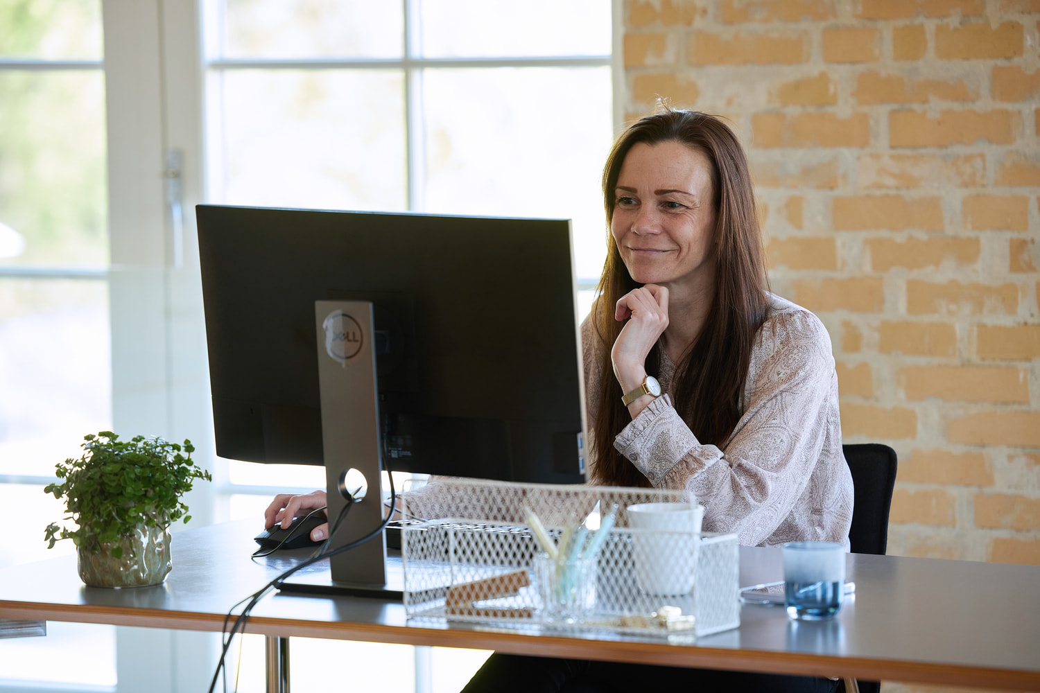 A woman is sitting at a desk with a computer and working while she is smiling.