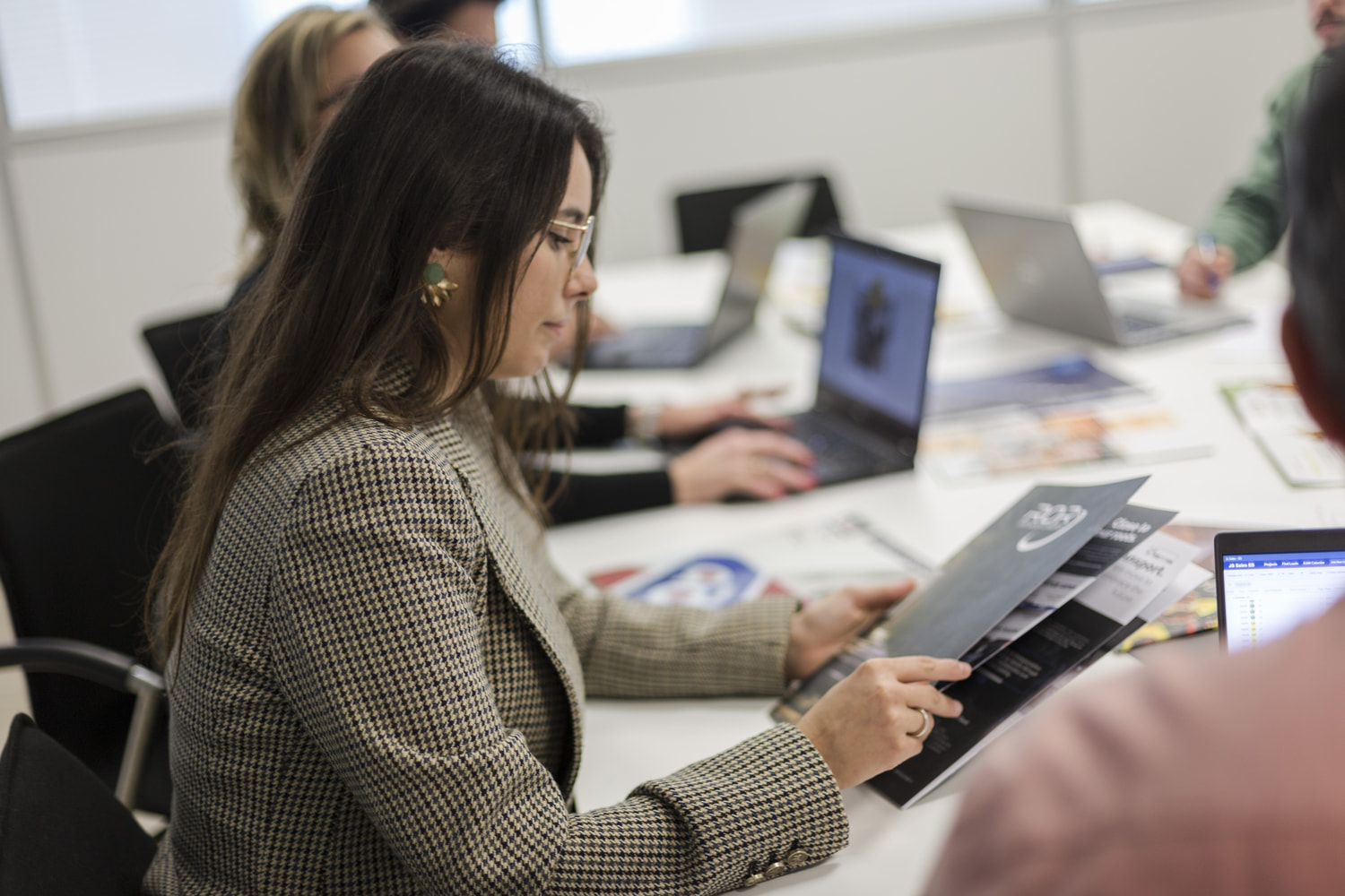 A woman is at a meeting with multiple colleagues. She is sitting at a desk and looking through a brochure.