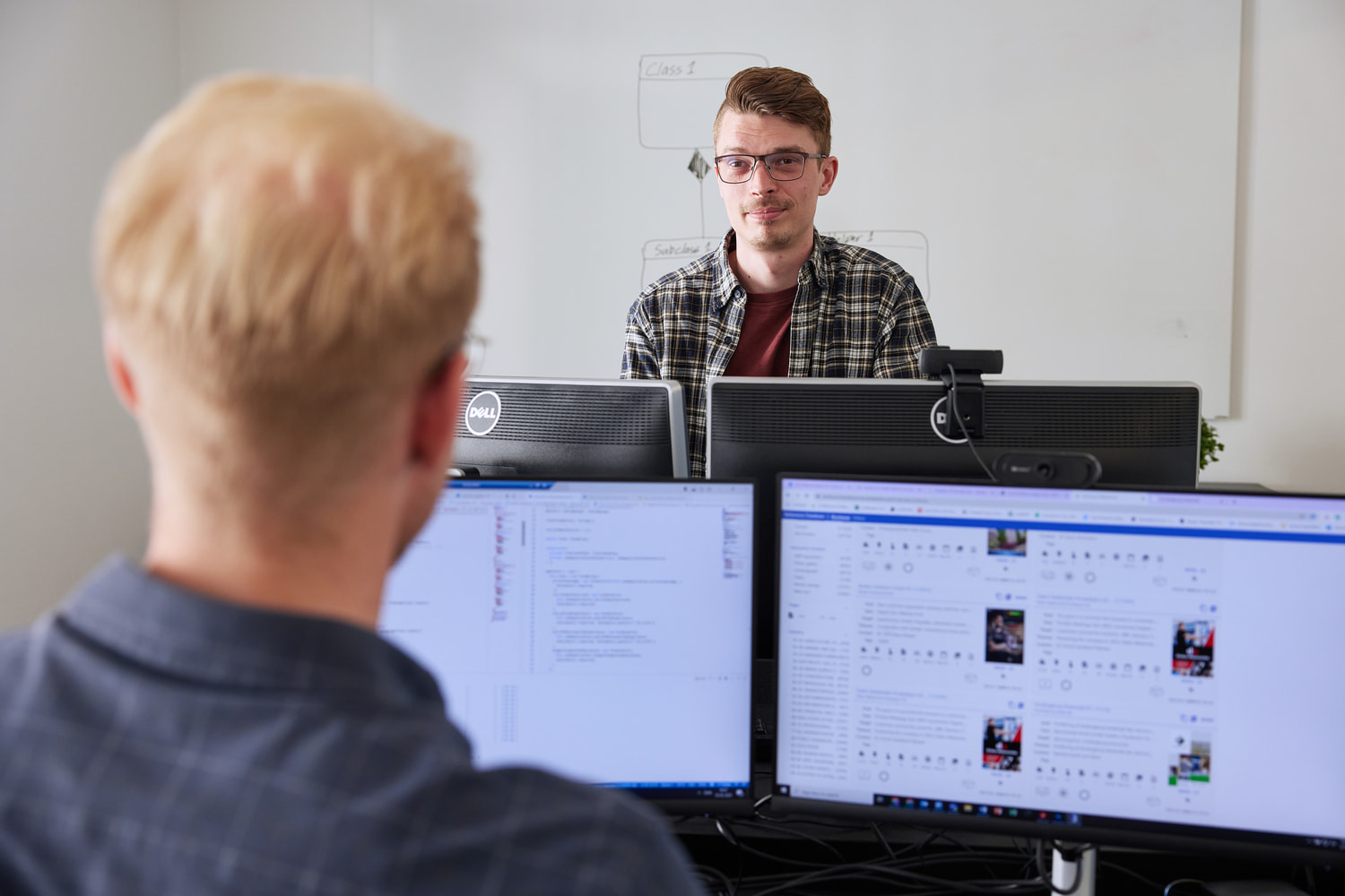 Two men sitting at desks across each other and talking. Viewing one of them from behind to see his screens.
